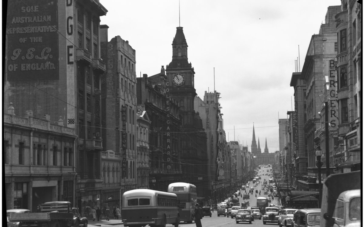Image of Bourke Street looking east; day time view photographed in the late 1940s or early 1950s. Two buses are prominent int he foreground, followed by a flatbed lorry. A large clock tower dominates the center of the image. A church steeple can be seen int he background.