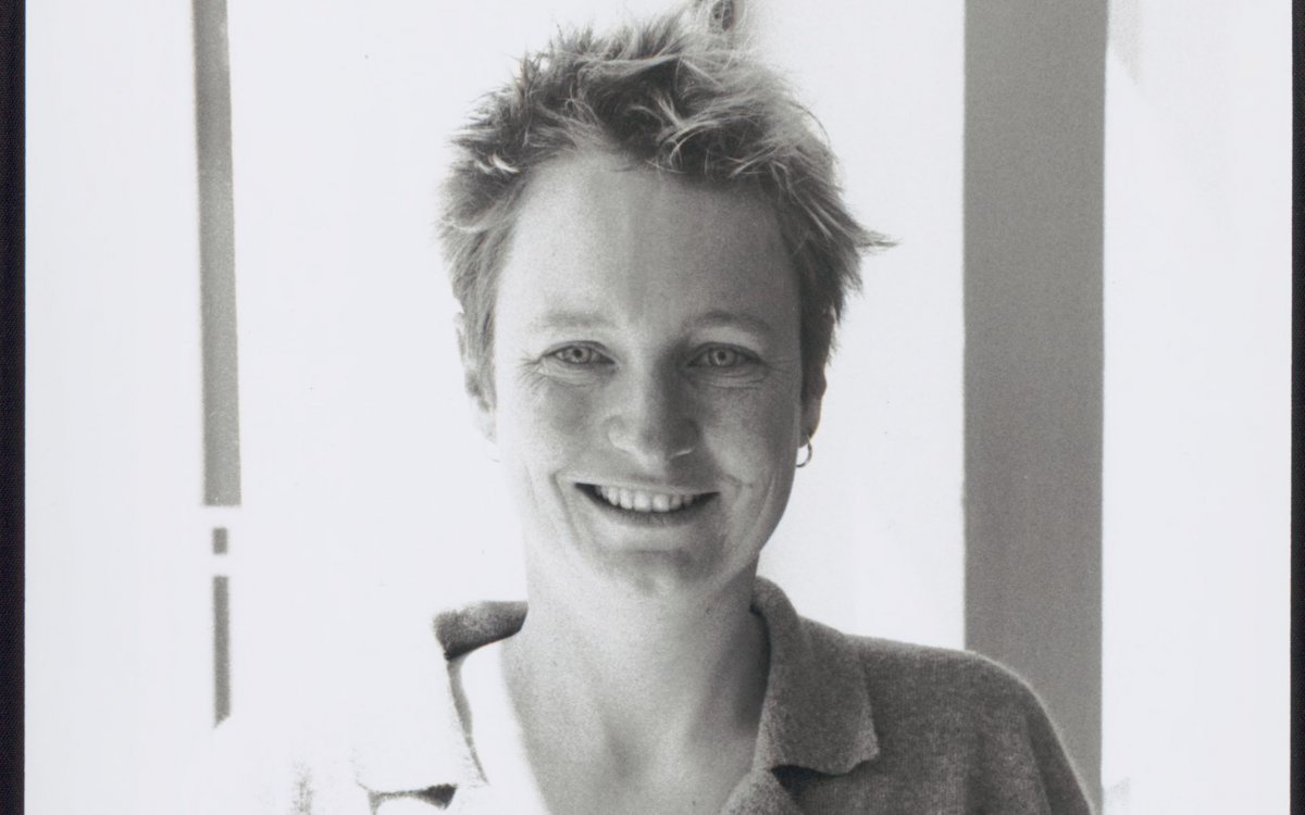 A black and white portrait photo of a young, freckle faced, smiling woman with very short, wild, fair hair, holding a book open. 