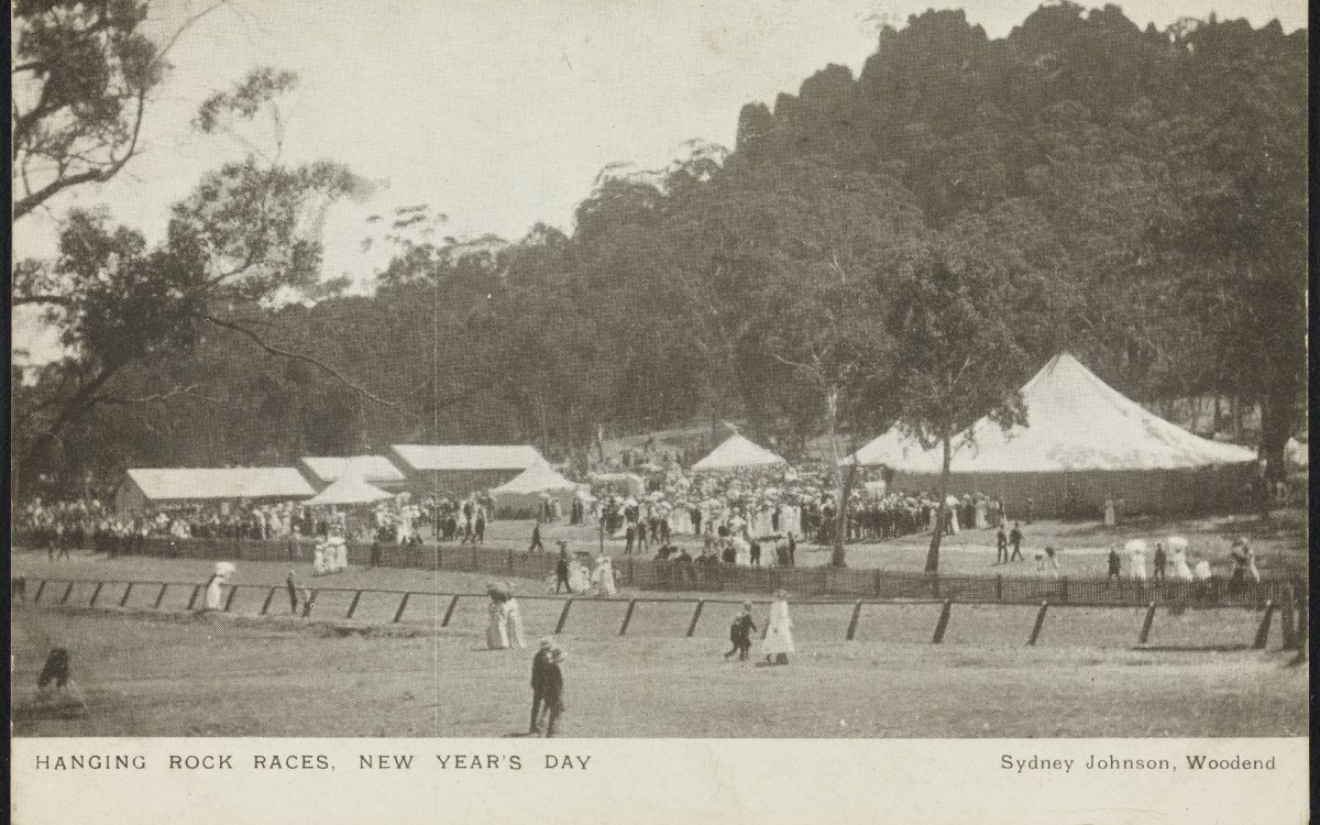 An old black and sepia toned photo of part of a race course, several tents and hundreds of people dressed in clothes from 1910, and a rocky topped mountain. 
